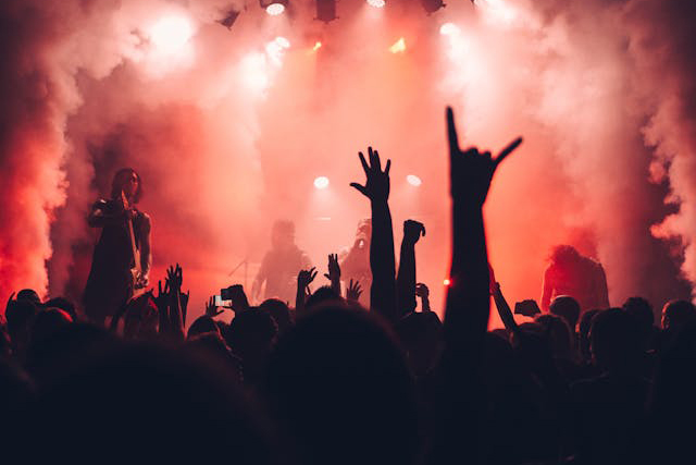 crowd in darkness in front of red-lit smoky stage, and raised in devil-horns sign